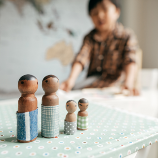 Small child sitting in the background. Four wooden pegs are in the foreground, two adults and two children.